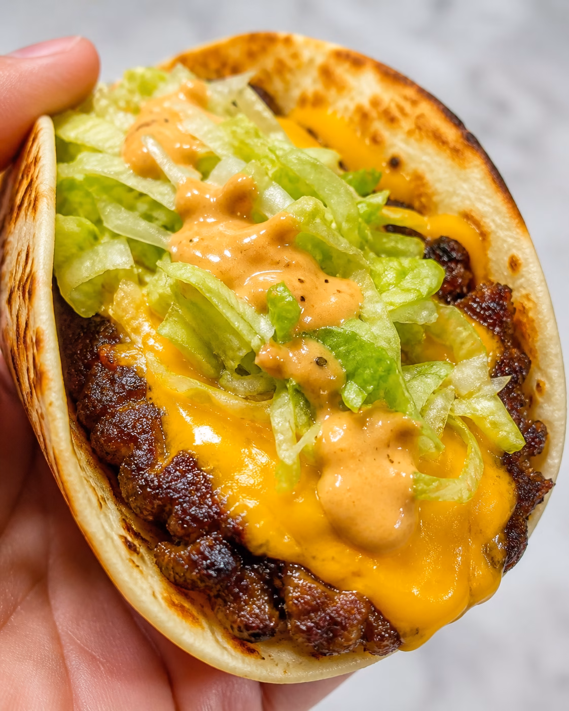 Close up of a crispy smashburger patty pressed onto a toasted flour tortilla, topped with perfectly melted American cheese, fresh shredded lettuce, and a creamy burger sauce, held in someone's hand.